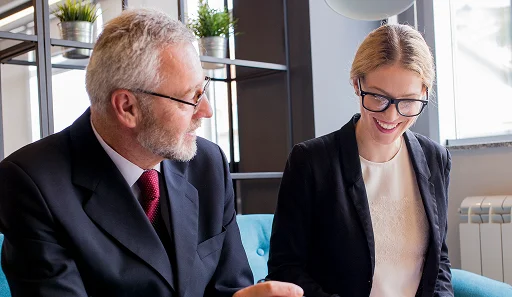 Mężczyzna i kobieta w strojach biznesowych siedzą na niebieskiej sofie i przeglądają dokumenty. Sceneria to jasne biuro z półkami i roślinami doniczkowymi, które nadają mu profesjonalny charakter