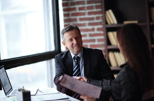 Mężczyzna i kobieta w strojach biznesowych podają sobie ręce nad biurkiem, na którym widoczne są laptop i dokumenty, co sugeruje udane spotkanie. Pomieszczenie ma ceglaną ścianę i półki z książkami