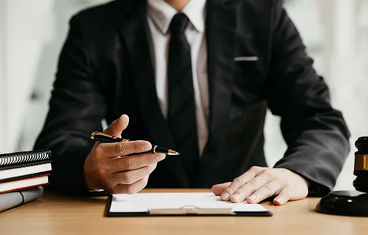 Osoba w czarnym garniturze siedzi przy biurku, trzymając pióro nad dokumentem, co sugeruje, że go przegląda lub podpisuje. W pobliżu znajdują się młotek i notesy