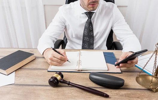 Osoba w czarnym garniturze siedzi przy biurku, trzymając pióro nad dokumentem, co sugeruje, że go przegląda lub podpisuje. W pobliżu znajdują się młotek i notesy